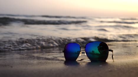 Sea-waves-washing-over-sunglasses-against-a-backdrop-of-a-setting-sun