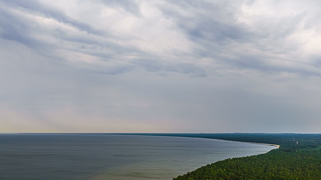 Overcast raining sky above calm bay and forested coastline, moody weather and muted colors, static timelapse