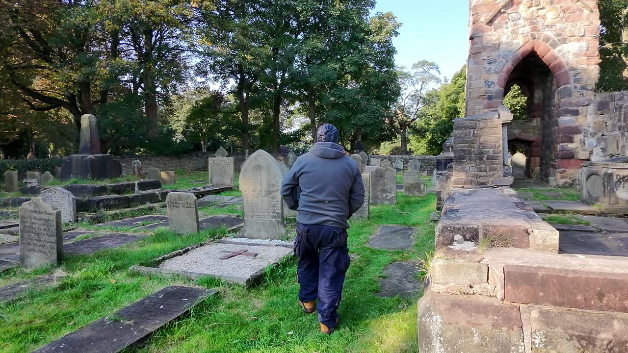 Following bearded male wearing hat and casual clothing walking in sunlit cemetery church ruins