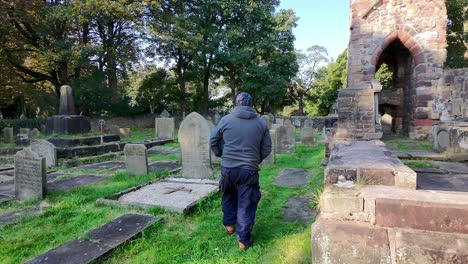 following bearded male wearing hat and casual clothing walking in sunlit cemetery church ruins