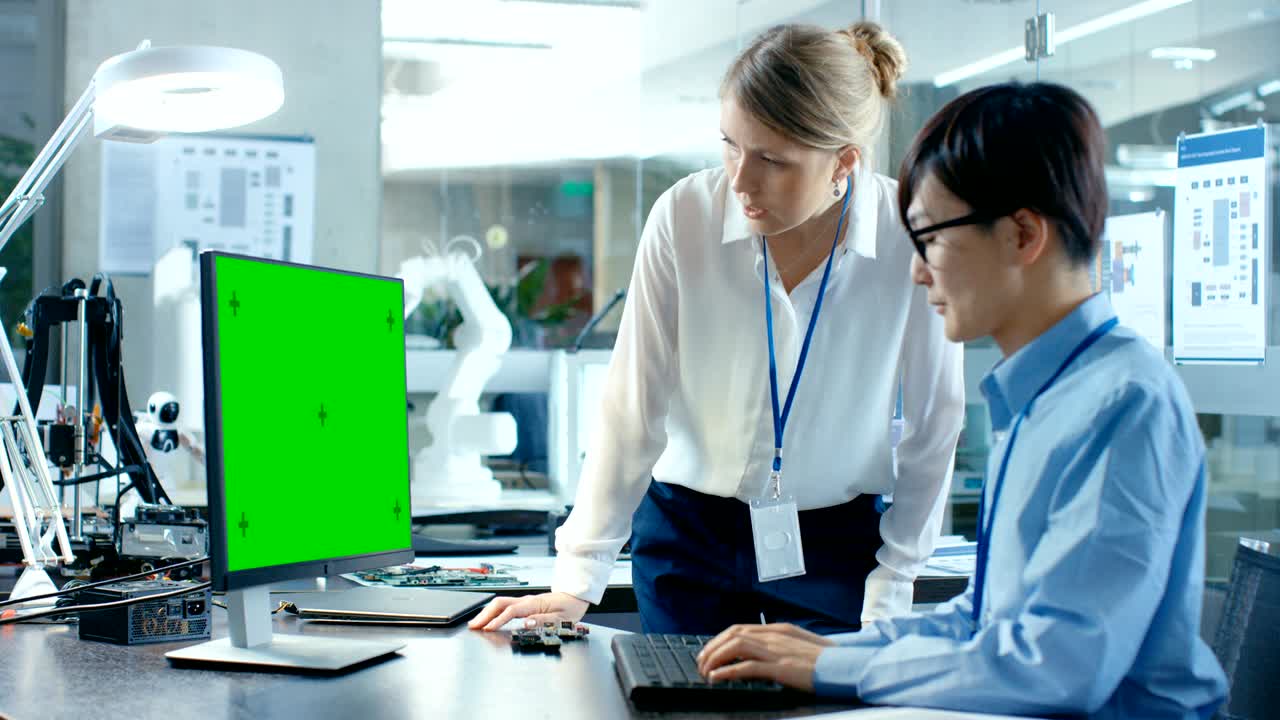 그의 책상에 앉아있는 아시아 과학자는 녹색 화면 개인 컴퓨터에서 일하는 동안 수석 엔지니어와 상담합니다. 배경에는 로 팔 모델과 함께 컴퓨터 과학 연구 실험실이 있습니다.
