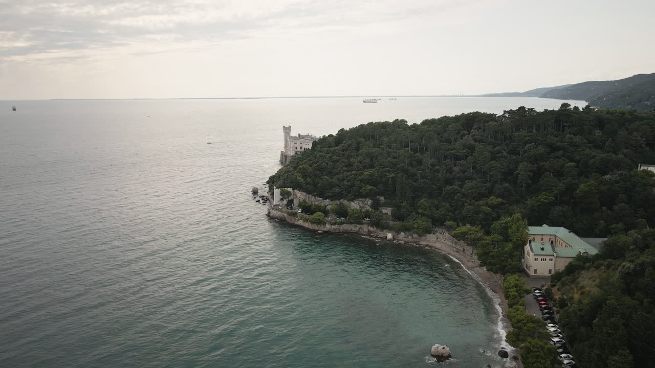 Un drone cinematico 4K UHD vola in avanti lungo la costa adriatica di Trieste verso il Castello di Miramare, rivelando un promontorio boscoso e un mare calmo per spot di viaggio, intro.