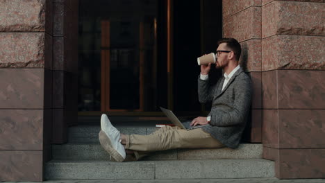 stylish businessman sitting outdoors on the steps of a building, drinking coffee and working on laptop computer