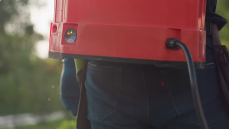 close up rear view farmer wearing mask and gloves carrying red tank sprayer fogging green crop leaves with fine mist under sunlight, blurred trees in background