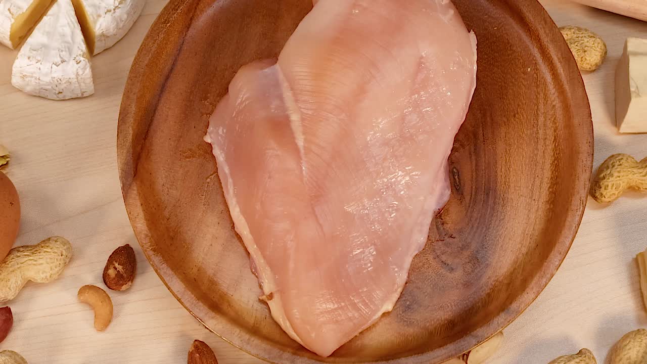 Close-up of chicken breast on a wooden plate surrounded by assorted nuts and cheese wedges.
