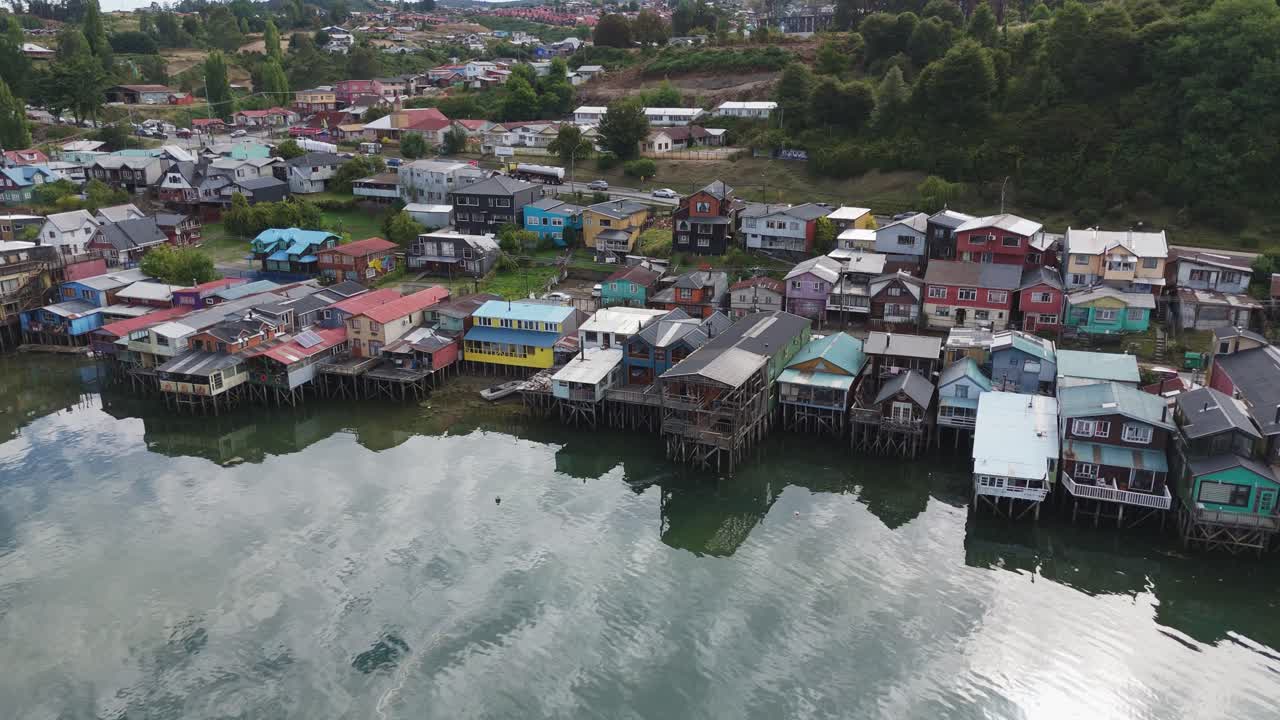 Vibrant stilt houses reflected on waterfront in Castro, Chiloé Island, surrounded by nature. Aerial Parallax Shot