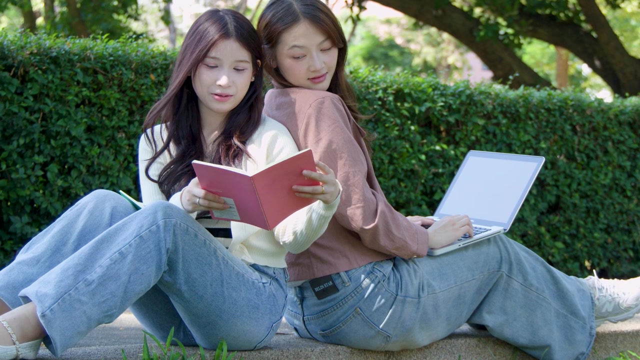 屋外で背中合わせに座り、一人はノートパソコンを使い、もう一人は読書をしている2人の女子学生