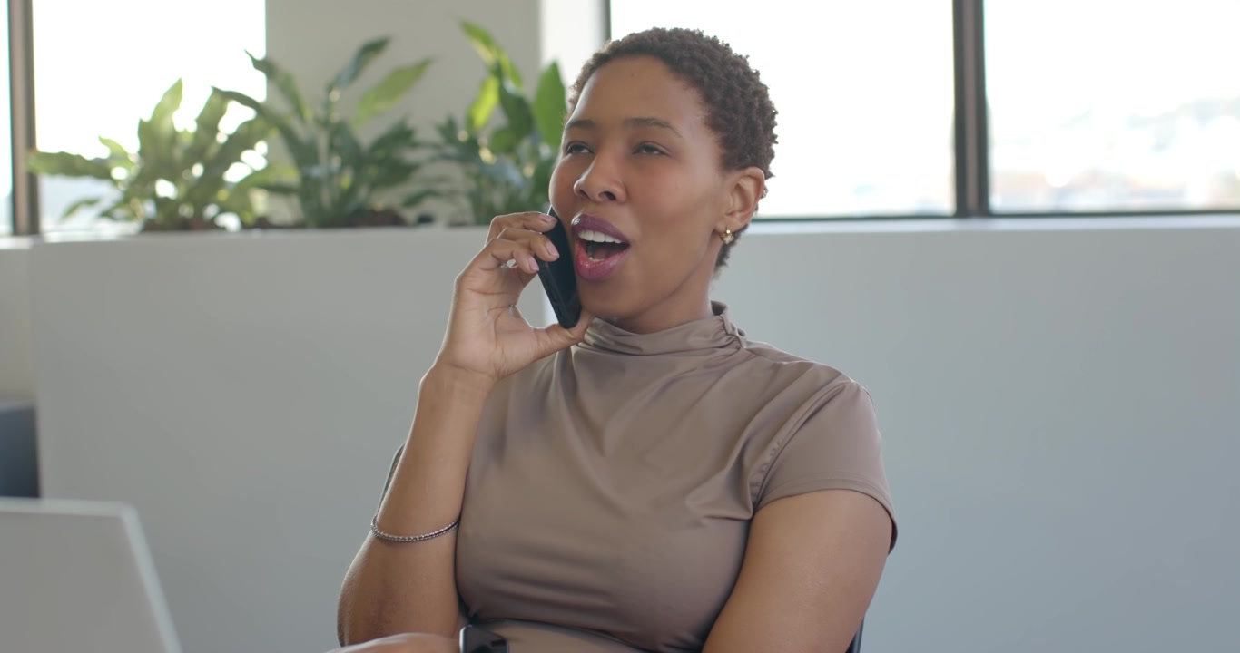 Mulher afro-americana recebendo uma chamada, segurando o telefone e falando a trabalho em uma mesa no escritório com um laptop.