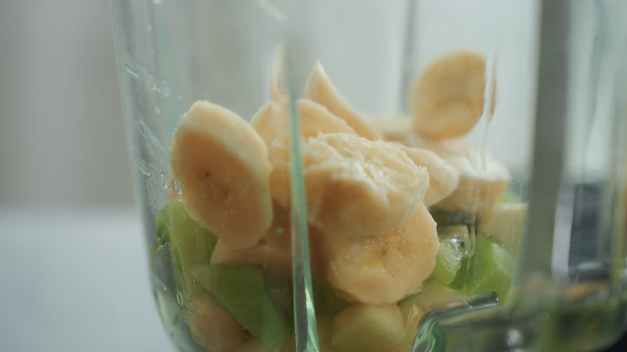 Banana e kiwi a fette nel frullatore, primo piano di un barattolo di vetro con strati di frutta fresca, luce naturale soffusa, polpa e umidità visibili, preparazione sul bancone della cucina per un frullato sano per la colazione, delicato.