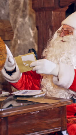 santa with a globe. mature man in red costume of santa sitting at the table in the room and looking at the globe. christmas time. vertical video