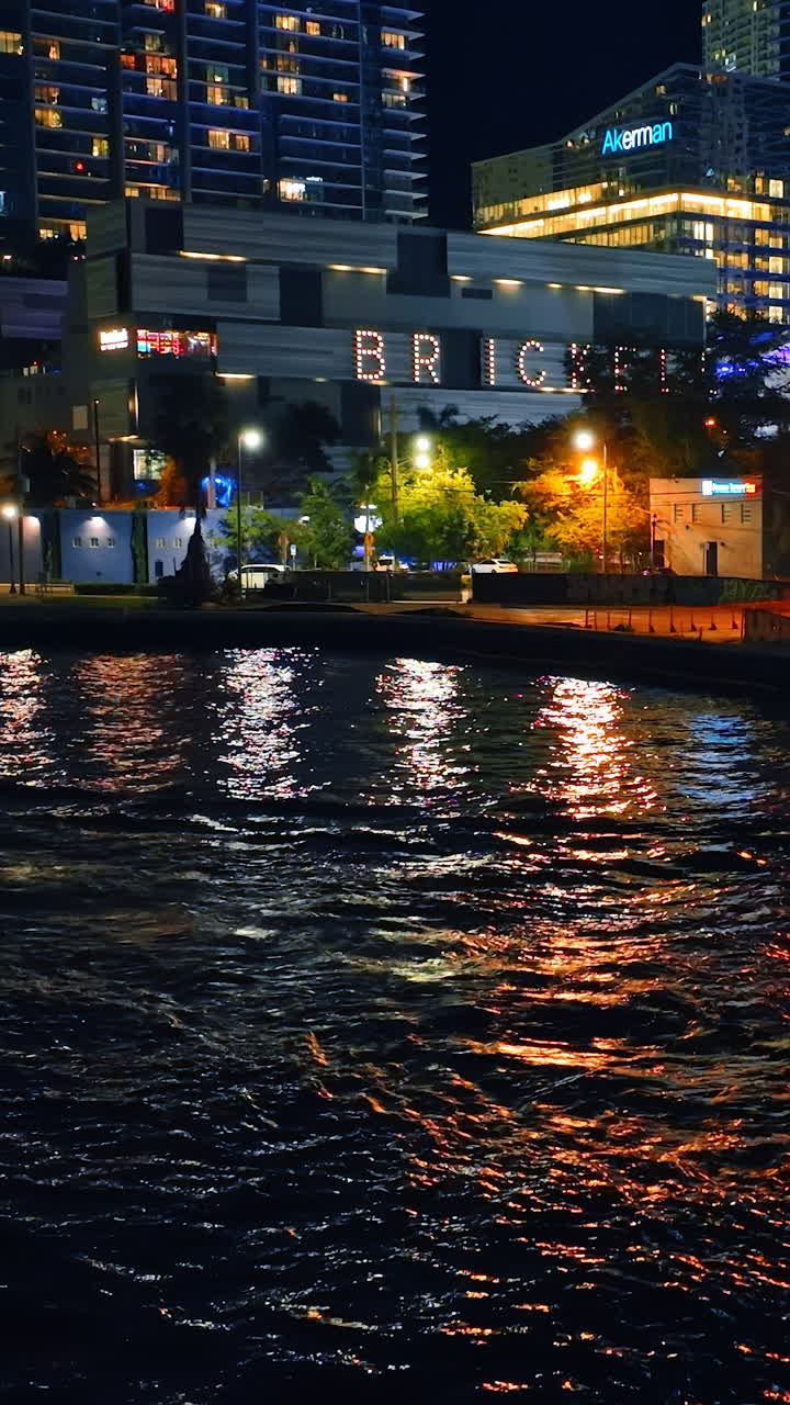 Motor boat going by the river at night. Buildings full of lights on the waterfront of Miami, Florida, USA. Vertical video