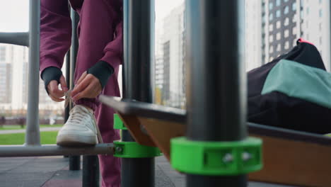 trainer approaches exercise bar carrying gym bag, sets bag at corner of equipment, bends gracefully to tie shoelace on bar before beginning workout under open sky in urban outdoor fitness area