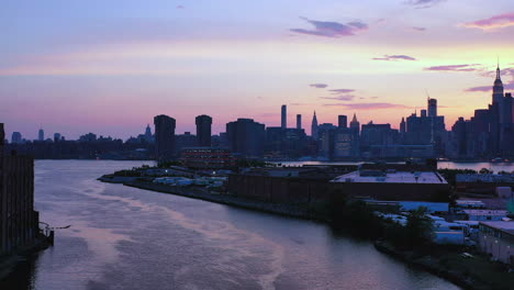 aerial drone footage pull away over purple water from the refection of the purple sky sunset, as camera pulls straight back from new york city skyline view