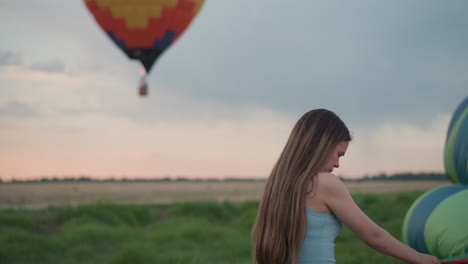 side view of young woman in denim shorts holding hot air balloon fabric near fan engine while colorful envelope floats blurred overhead against pastel sunset sky above grassy field