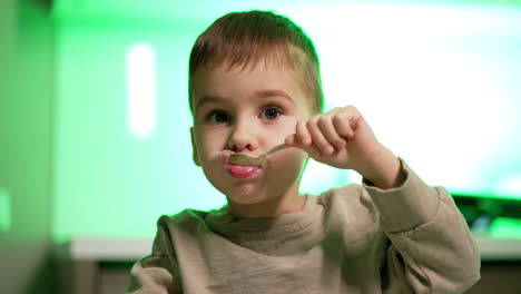beautiful toddler boy holding a spoon in his mouth. adorable kid eats yogurt and smiles to camera. close up. blurred backdrop.