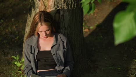 lady sits under tree reading book while leaning against trunk, she rubs her eyebrow and adjusts her hair, appearing thoughtful, sunlight softly illuminates her face, with leaves swaying by the wind