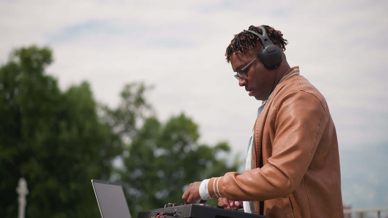 black DJ rooftop mixing with headphones soulful street musician adjusting controller against cloudy sky brown jacket and glasses intimate outdoor setup focused beatmatching mellow groove urban park