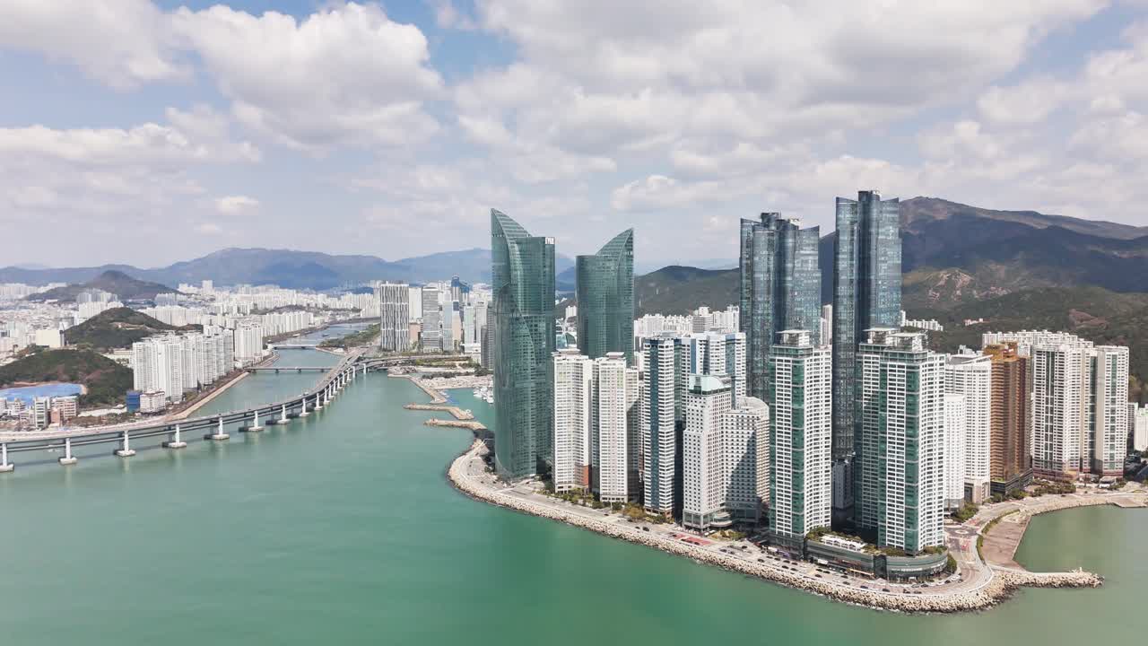 Aerial perspective of Busan, South Korea highlighting modern skyscrapers, waterfront towers, turquoise sea, a long coastal bridge, and surrounding mountains combining urban growth with natural scenery