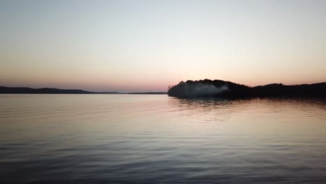 a calm lake wisconsin reflects the pastel tones of twilight as smoke drifts from a wooded peninsula. this wide aerial view captures serene beauty and atmospheric light in a peaceful dusk setting.