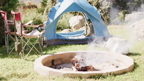 smoking firepit, tent, chairs and camping equipment in sunny countryside, copy space, slow motion