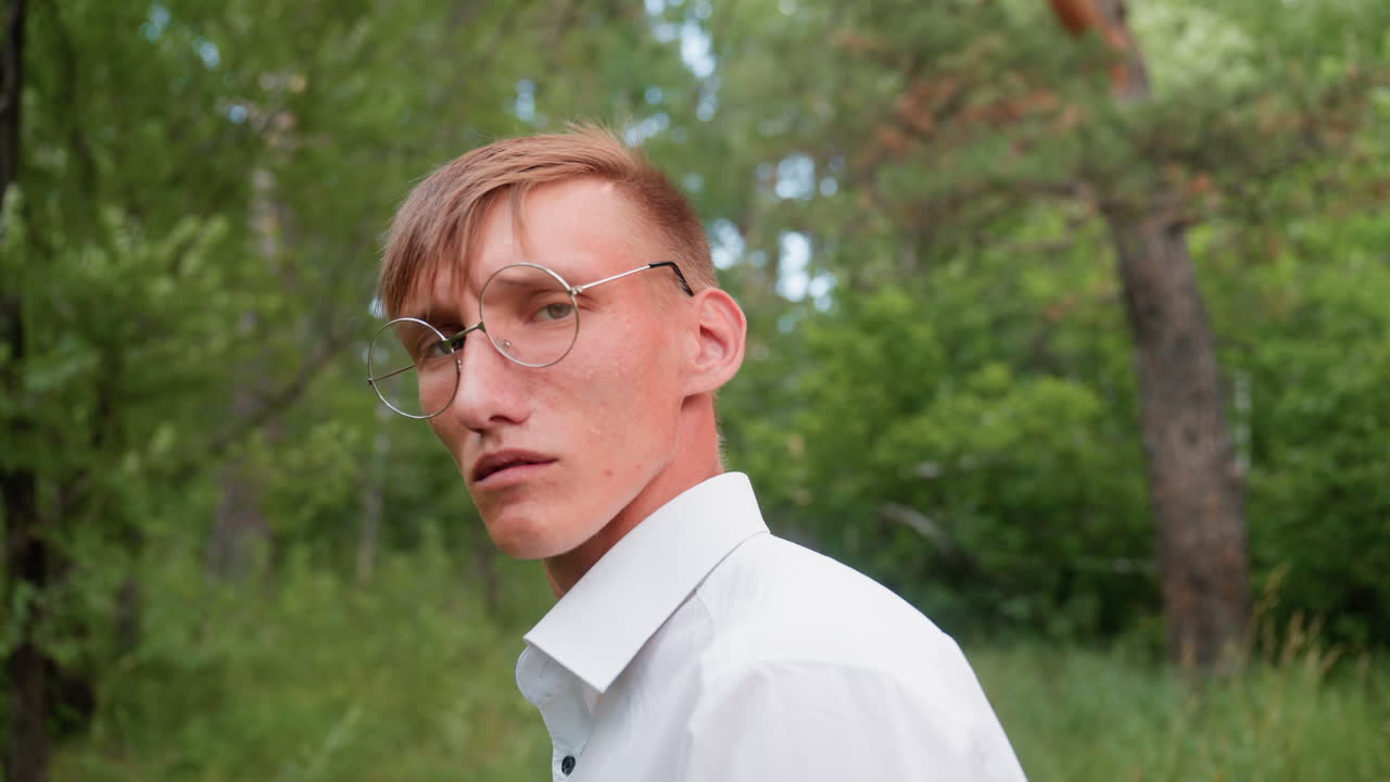 Back close view of handsome man wearing white shirt and glasses turned back with frown looking at trees in forest under daylight surrounded by greenery expressing curiosity and thoughtful observation