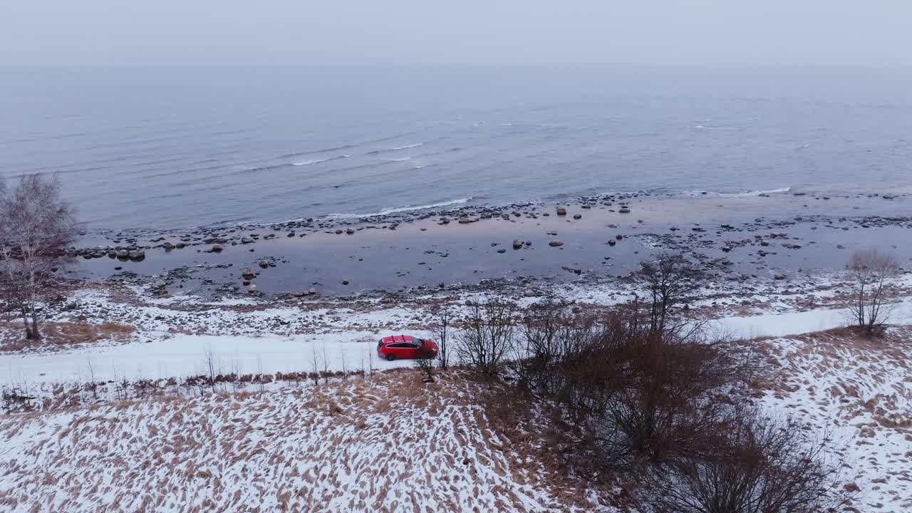 Baltic Sea horizon fades in fog as red car travels snowy seaside path near Tūja