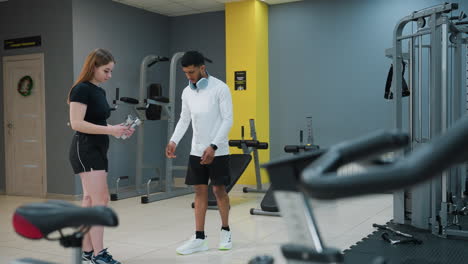 trainer in white sportswear hands dumbbell to female client during indoor gym session, gently places hand on her shoulder while guiding her through stamina-building exercise surrounded by equipment