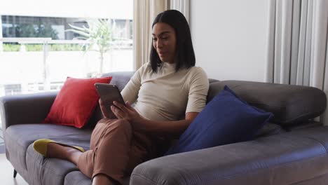 Mixed-race-gender-fluid-person-sitting-on-couch-and-using-tablet-at-home