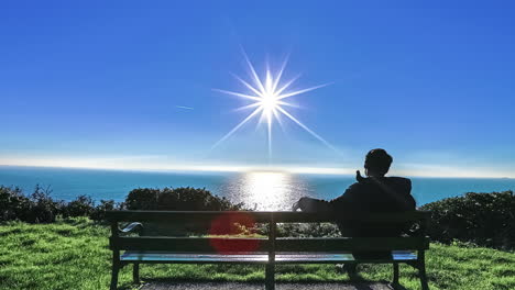 Joven-En-Un-Banco-Del-Parque-Mirando-El-Océano-Con-El-Sol-Reflejándose-En-El-Agua---Lapso-De-Tiempo-De-Revestimiento