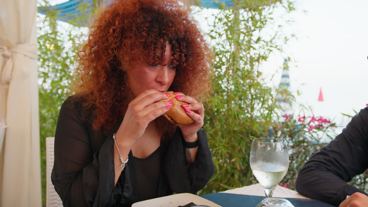 Curly Woman Biting The Burger At The Restaurant On The Beach For Lunch