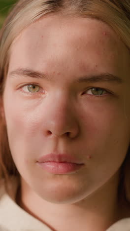 close-up of young lady with intense expression, sunlight reflecting softly on her face, adding warmth and depth to her look, with blurred green foliage in background
