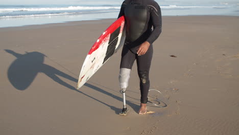 carrellata di un surfista maschio con gamba artificiale che cammina lungo la spiaggia e tiene la tavola da surf sotto il braccio