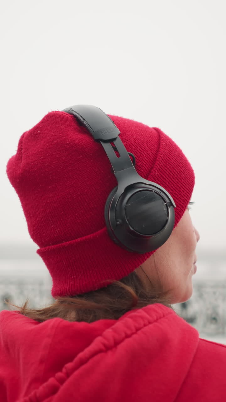 vista trasera de una mujer con gorra roja y sudadera con capucha escuchando auriculares, mirando hacia el lejano horizonte nebuloso durante el invierno, el fondo presenta una valla decorativa y un ambiente nebuloso atmosférico