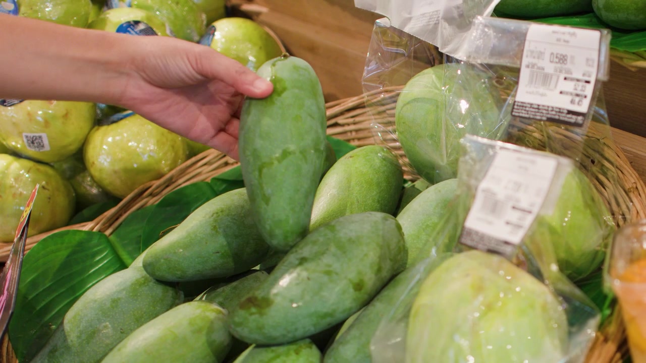 La mano di una persona seleziona e ispeziona manghi verdi freschi da un cesto in un supermercato.