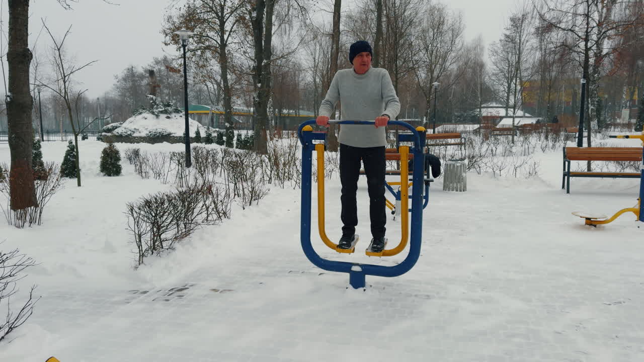 Aged Caucasian male stands on the simulator in winter outdoors. Man exercising in the park on snowy day.