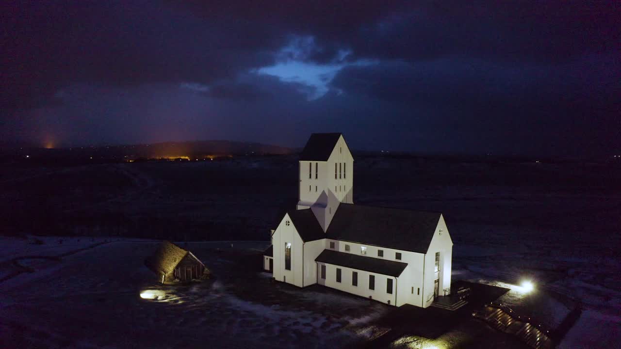 Overhead drone view of a remote Icelandic church at night, highlighting Nordic architecture, soft lighting, and the stillness of the surrounding rural town.