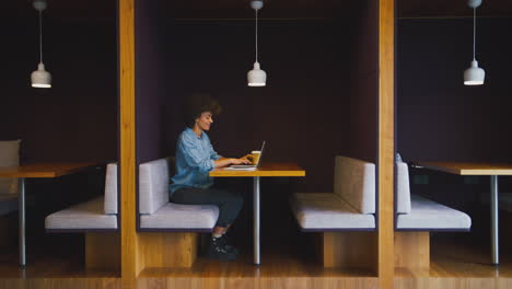 young businesswoman in modern office working on laptop in seating pod with takeaway drink