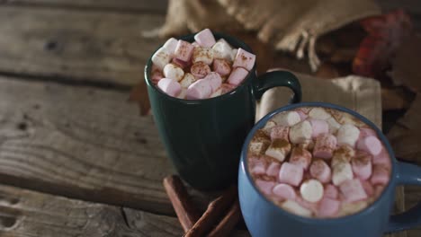 two cups of hot chocolate with marshmallows, autumn leaves and cinnamon sticks on wooden surface