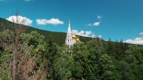 Iglesia-De-La-Natividad-Con-Cúpulas-Doradas-En-Shipka,-Bulgaria-Rodeada-De-Un-Denso-Follaje-Verde-Y-Exuberante,-Tiro-Aéreo-Inclinado-Hacia-Abajo