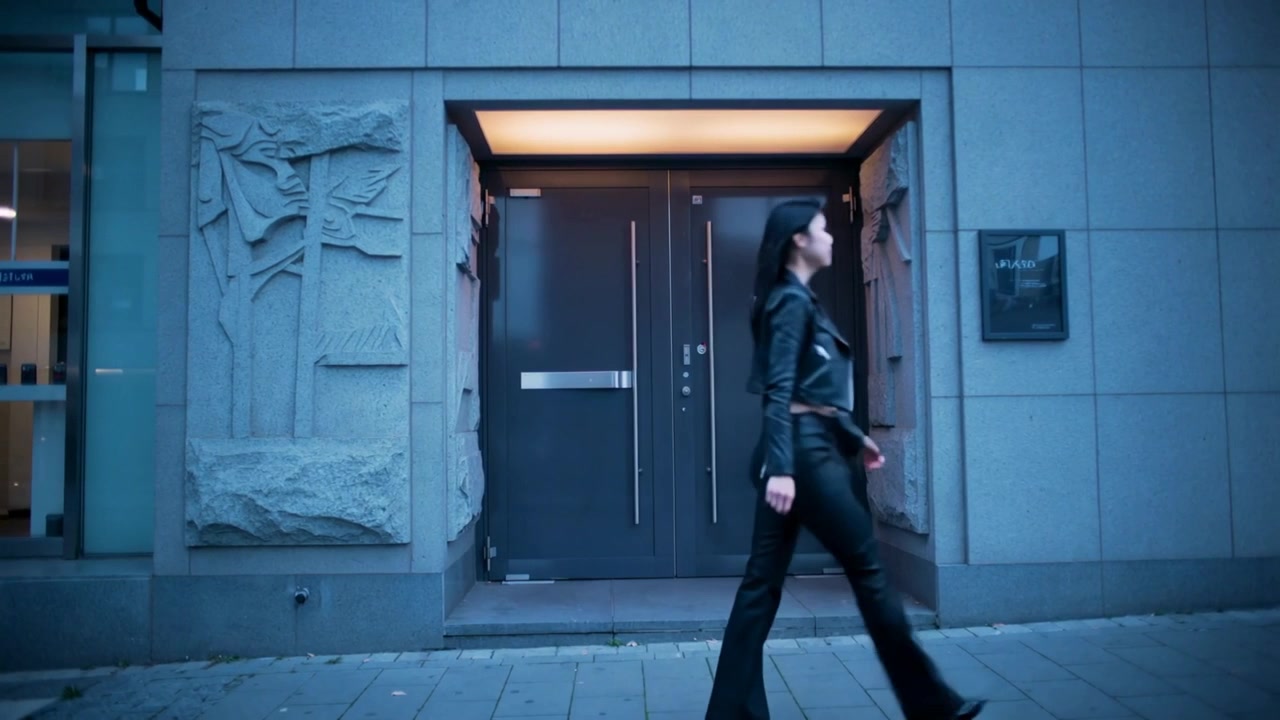 Entering frame, woman in leather jacket walking past recessed doors toward right under lit canopy
