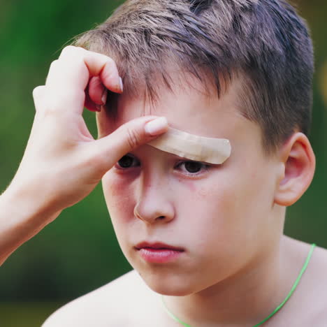 sad boy with injury on his eyebrow. woman's hands putting small white adhesive plaster on a wound outdoors. close-up.