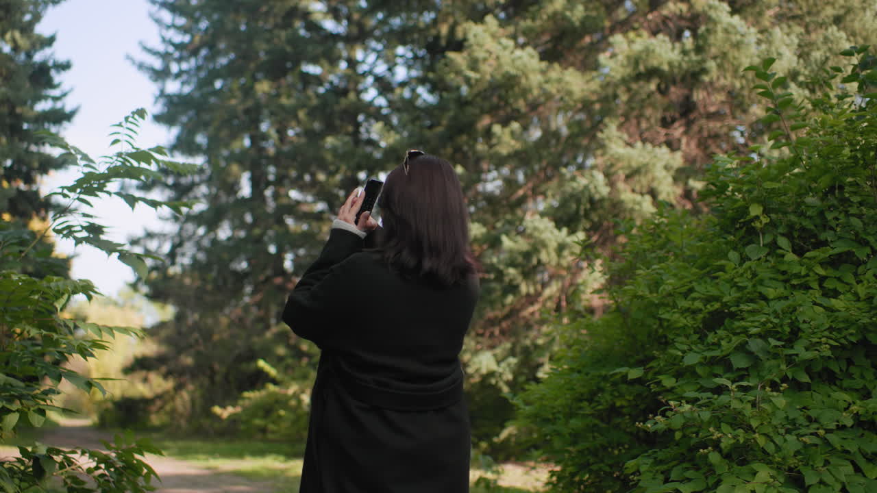 긴 겨울 코트를 입고 수목원을 거닐며 휴대폰을 들고 사진을 찍는 자연을 사랑하는 사람의 뒷모습, 상록수들이 길을 에워싸고, 부드러운 햇살, 평화로운 산책 기록
