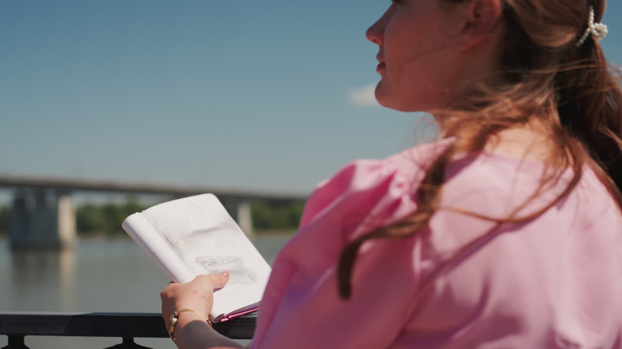 Giovane donna bianca che legge un libro in riva al fiume, capelli intrecciati illuminati dal sole, vestito rosa, ringhiera in metallo decorata, vista rilassante sul fiume, ponte in lontananza, studentessa che sfoglia le pagine, sorriso pensieroso, brezza leggera, rilassata