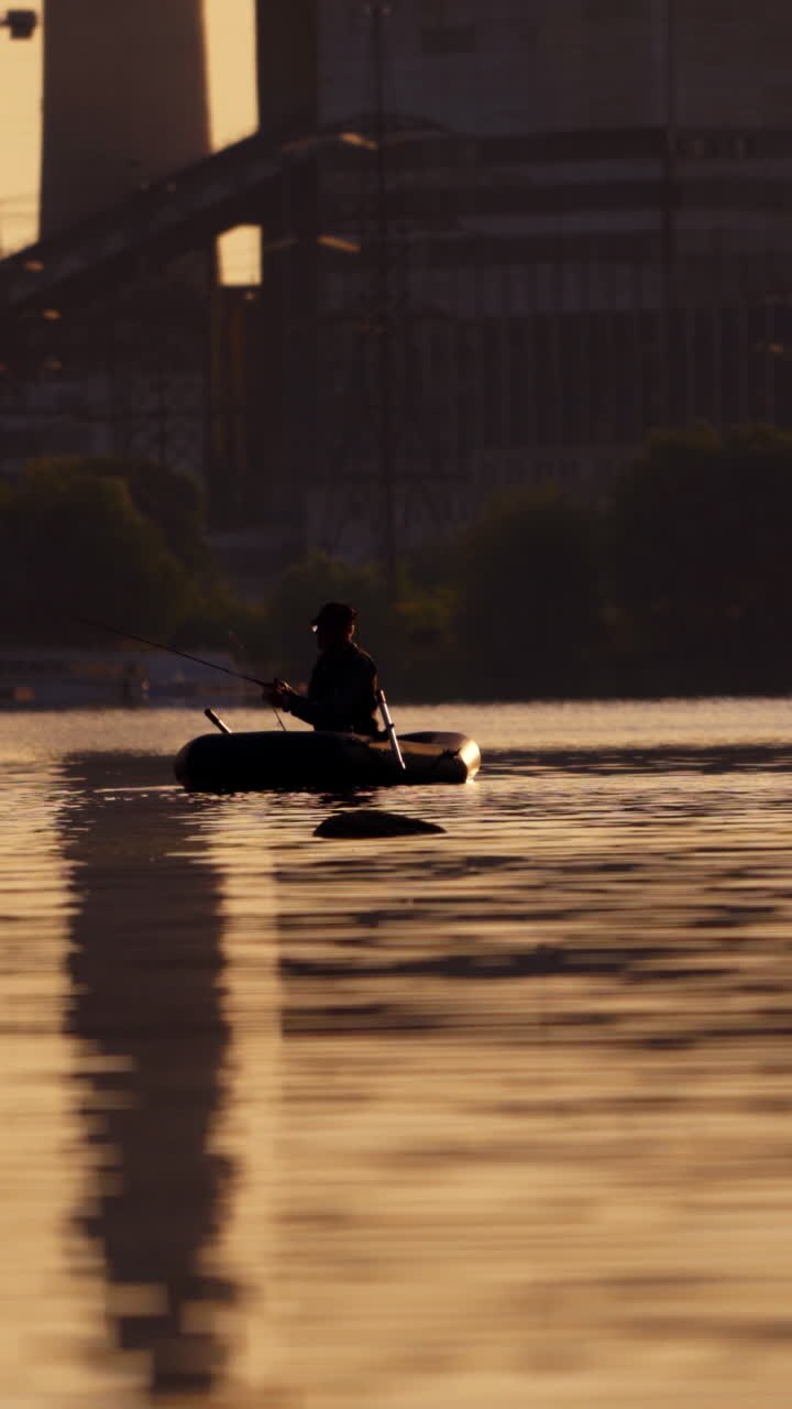 Fisherman in a boat with fishing rods in the evening. Man in hat floating in a boat and fishing on the background of a factory in the city. Vertical video