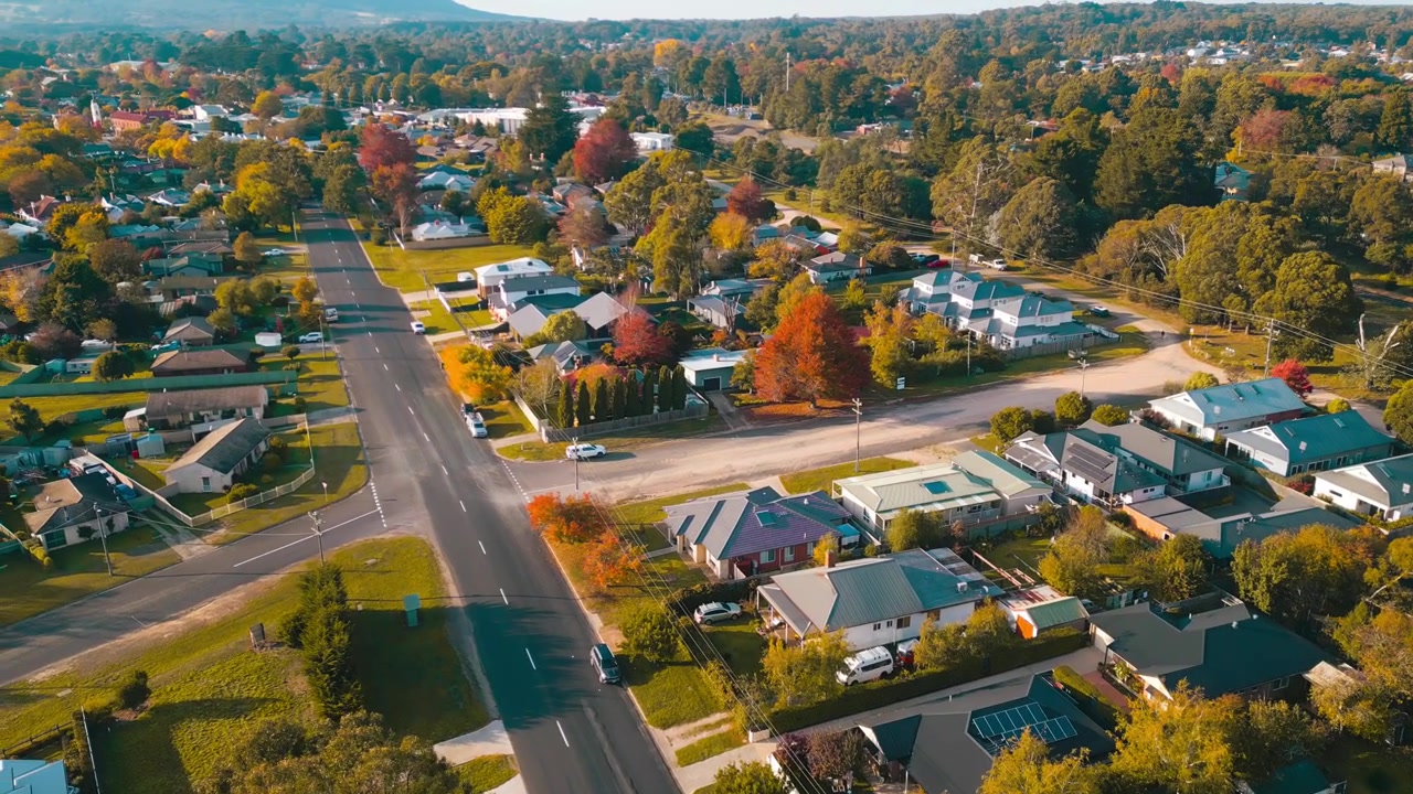 Filmato cinematografico con drone di case residenziali a Woodend, Victoria, con un movimento di panning verso l'alto e una leggera rotazione che rivela più del paesaggio suburbano, alberi autunnali e auto in movimento in una mattinata di sole