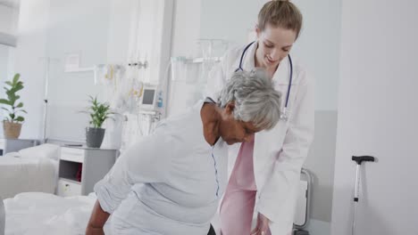Happy-diverse-female-doctor-helping-senior-female-patient-stand-from-bed,-slow-motion