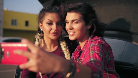 Vista-De-Cerca-De-Dos-Atractivas-Mujeres-Jóvenes-Posando-Mientras-Se-Toman-Selfie-En-El-Teléfono-Inteligente-Sentado-Dentro-Del-Maletero-Abierto