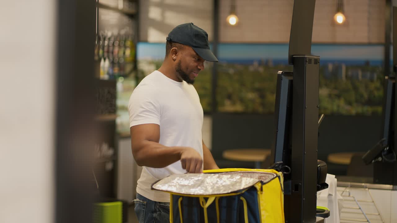 um homem de entrega de comida preto vestindo uma camiseta branca e um boné preto coloca as compras que ele comprou no caixa do supermercado em seu grande saco de carga de comida amarela