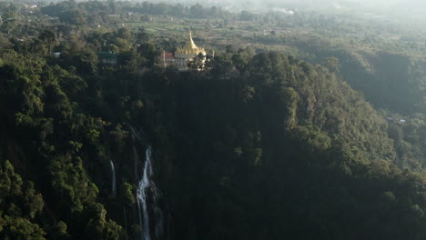 aerial backwards shot of golden buddhist temple and steep cliff with dat taw gyaint waterfall in myanmar