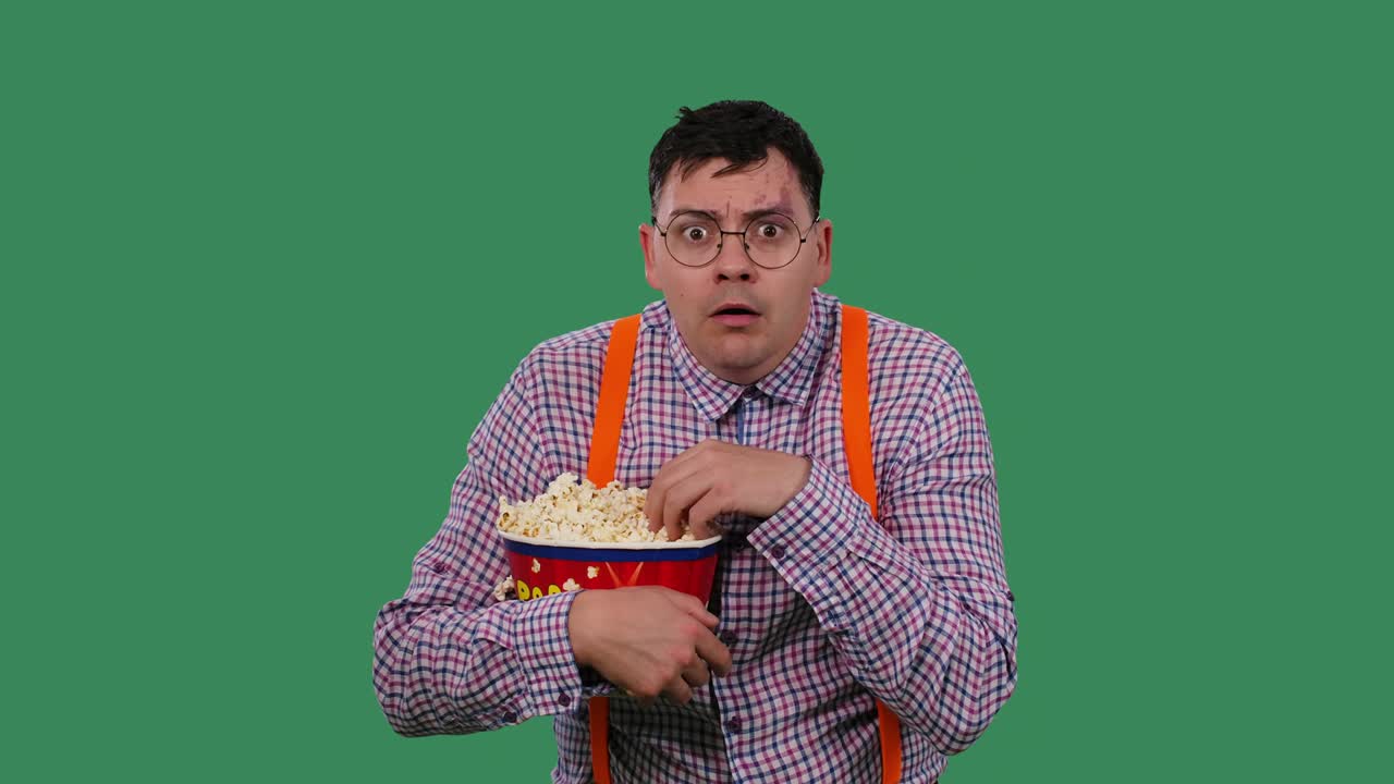 한 남자가 영화를 조심스럽게 보고 큰 바구니에서 팝콘을 먹는다. 초록색 스크린에 있는 스튜디오에서 오렌지색 서펜서를 입은 셔츠를 입고 안경을 쓴 남자의 초상화.